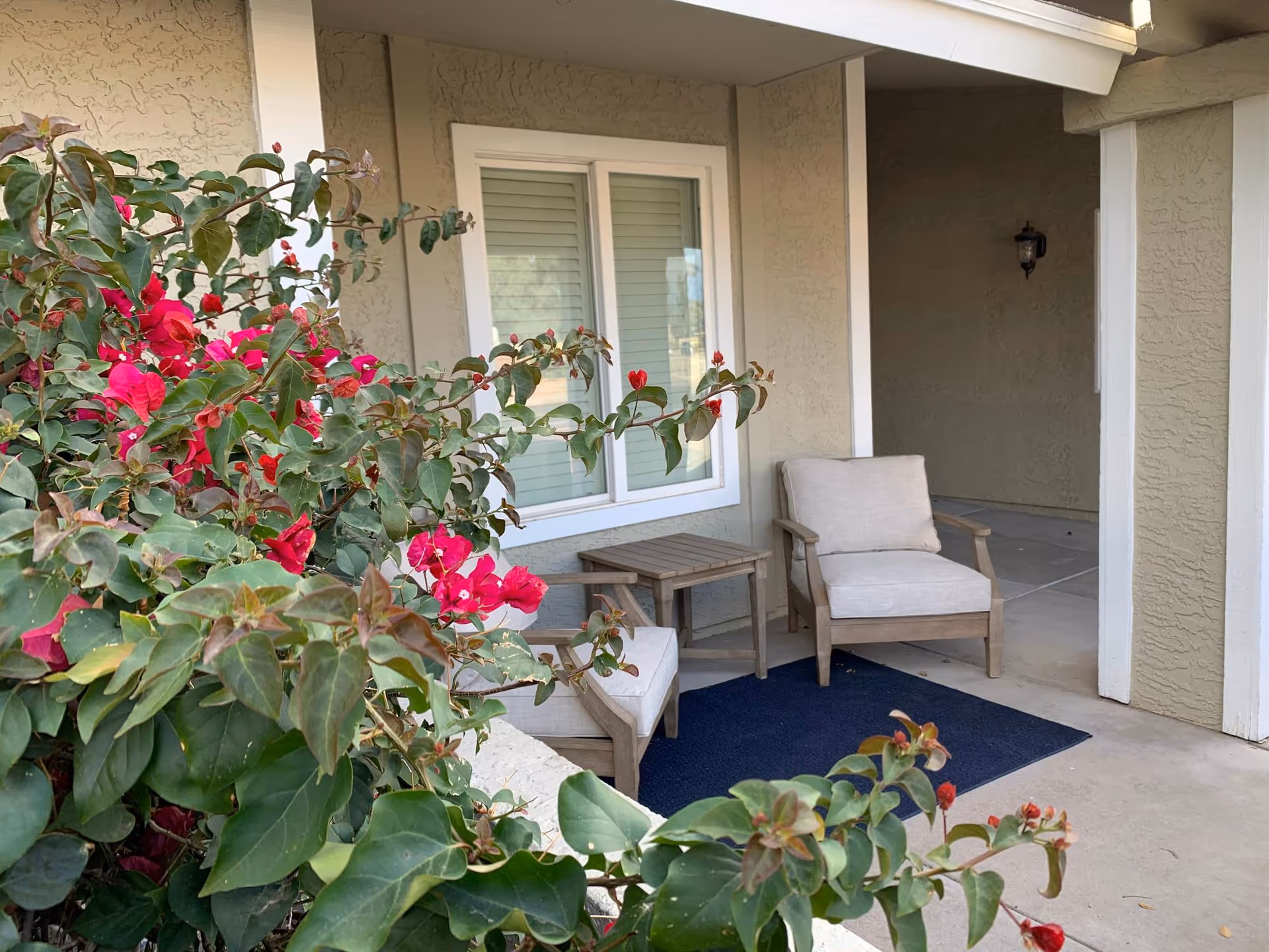 Outdoor front porch with beige chairs, small table, rug, and pink flowering bushes.