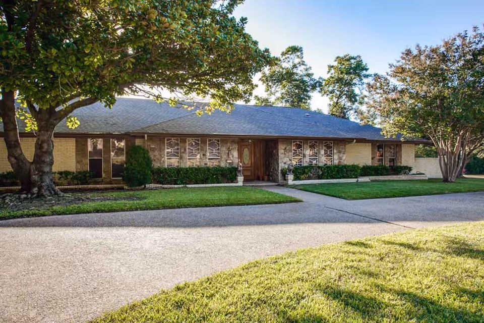 Single-story building with a gray shingled roof, stone and brick exterior, and multiple windows with decorative grills. The entrance features a wooden door with glass panels, flanked by two white planters. The building is surrounded by well-maintained green lawns, shrubs, and large trees providing shade.