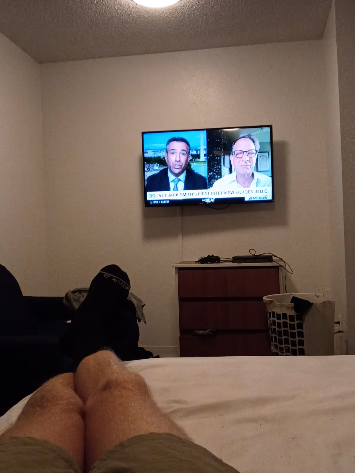 Person's legs visible on a bed in a small bedroom with a wall-mounted TV and dresser