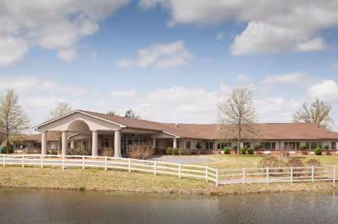 Single-story nursing facility with a covered entrance, white fencing, and lakeside grounds.