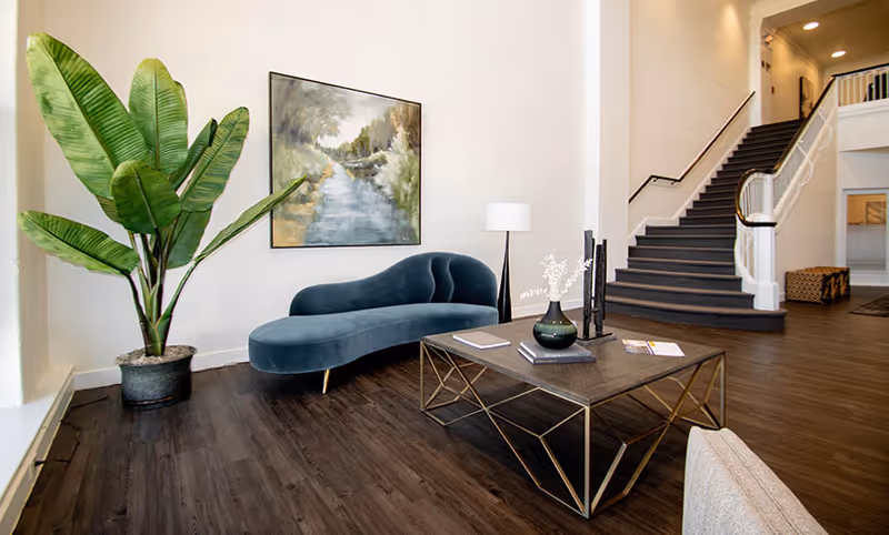 Stylish lobby seating area with a curved blue sofa, coffee table, large potted plant, wall art, and a staircase.