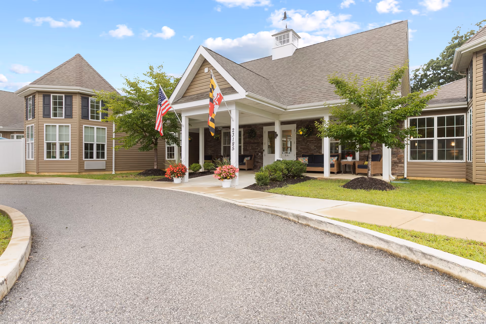 Exterior view of Discovery Commons At Wildewood senior living facility showing the main entrance with a covered porch, two flags, potted flowers, and surrounding landscaping under a partly cloudy sky.