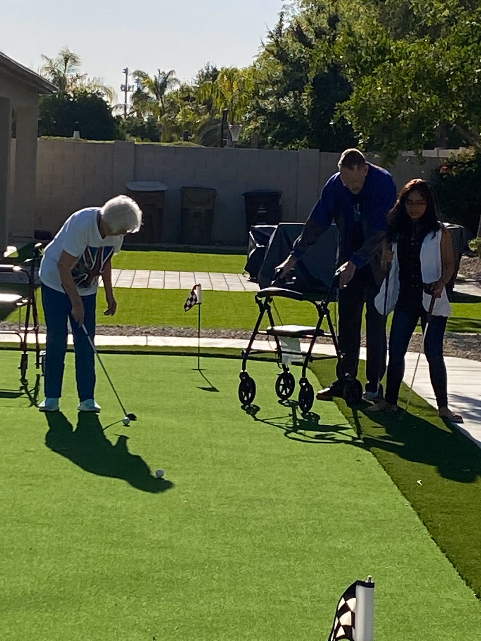 Elderly woman and companions play golf on a sunny outdoor putting green.