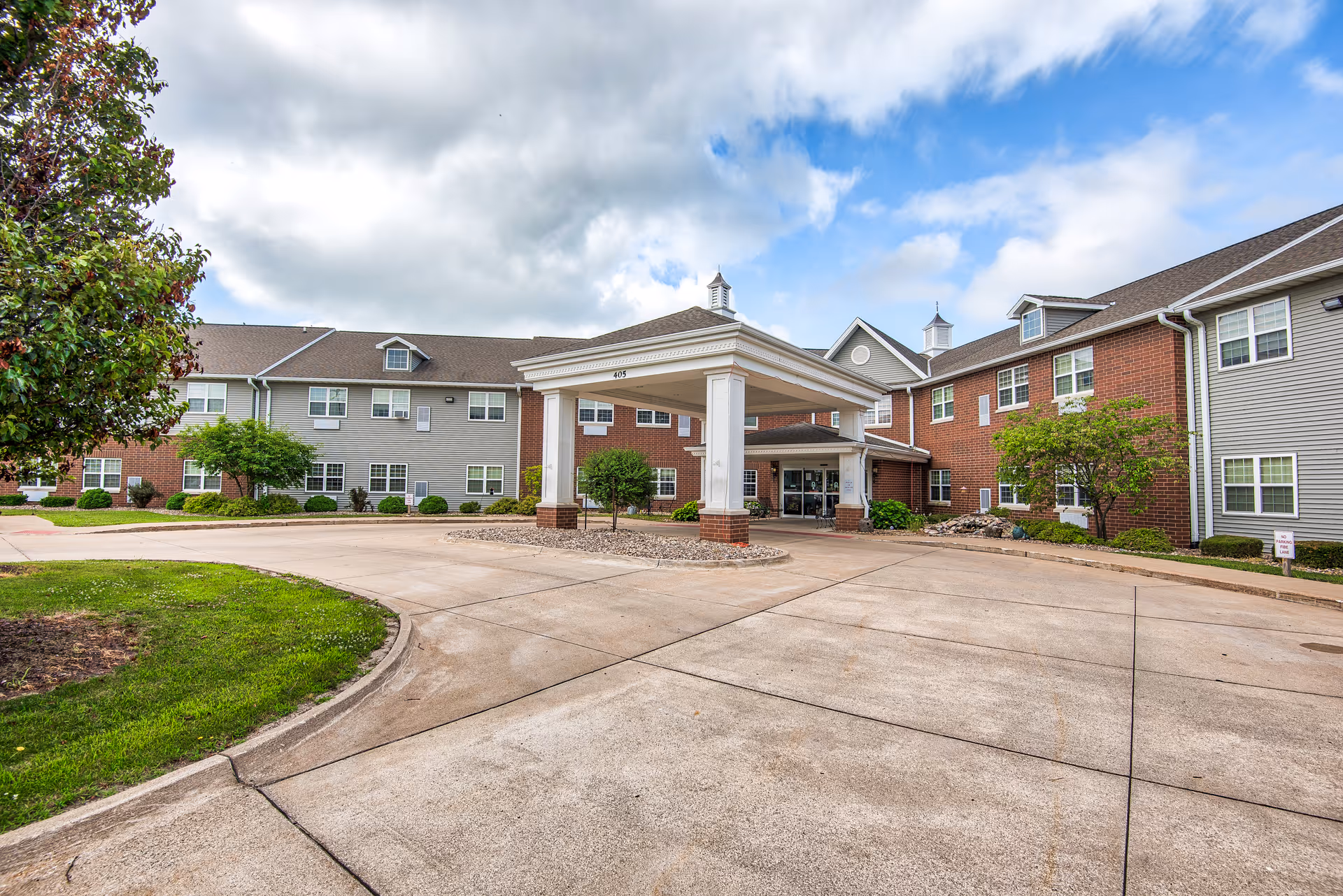 Front entrance of a two-story brick-and-siding senior living building with a covered porte-cochère and circular driveway.