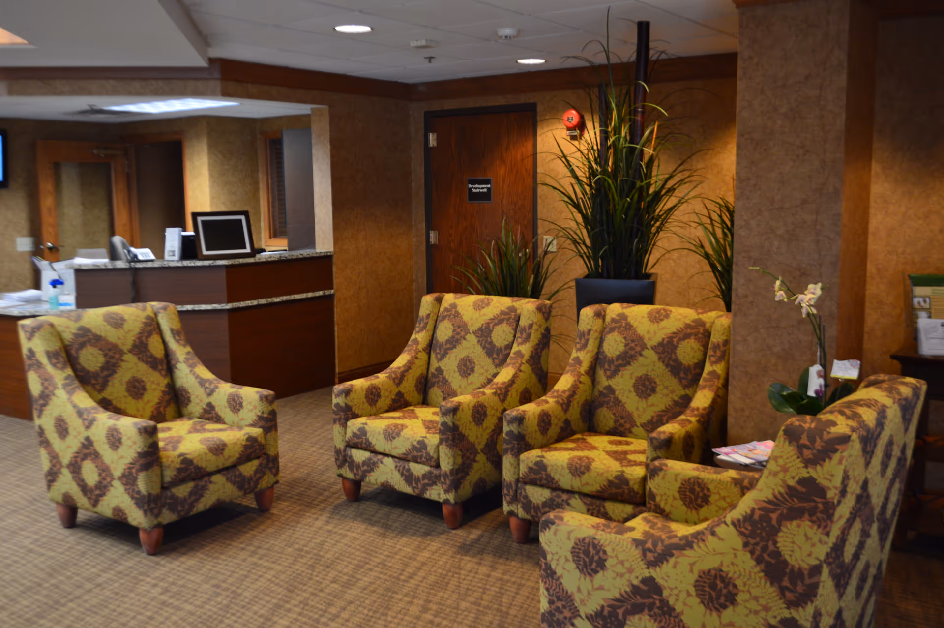 A seating area in a senior living facility with four patterned armchairs arranged in a semi-circle on a carpeted floor. Behind the chairs is a reception desk with a computer monitor and some paperwork. There are tall green plants in pots near a wooden door, and the walls are covered with brown textured wallpaper.