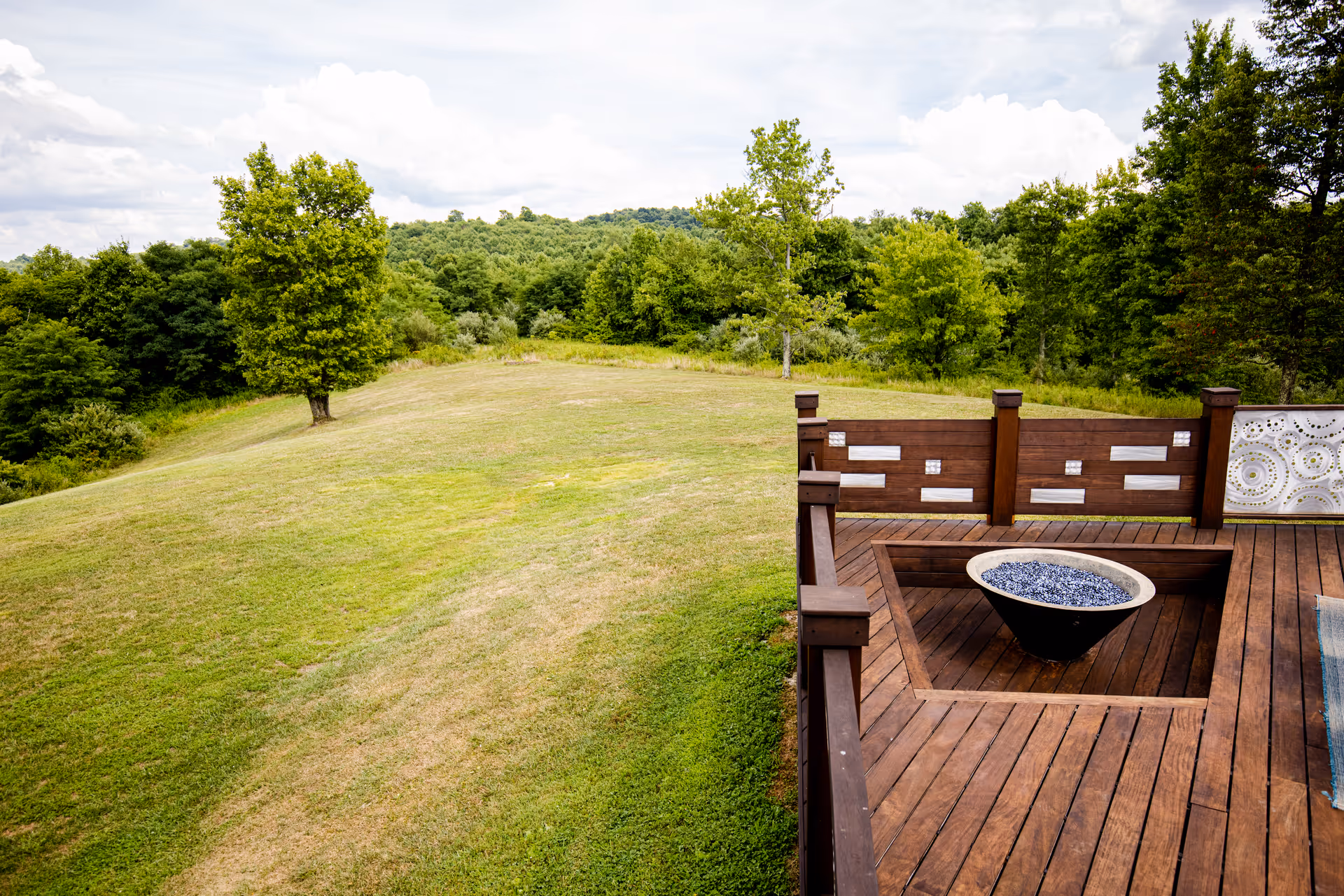 Wooden outdoor deck with a central fire pit overlooking a grassy, tree-filled landscape.
