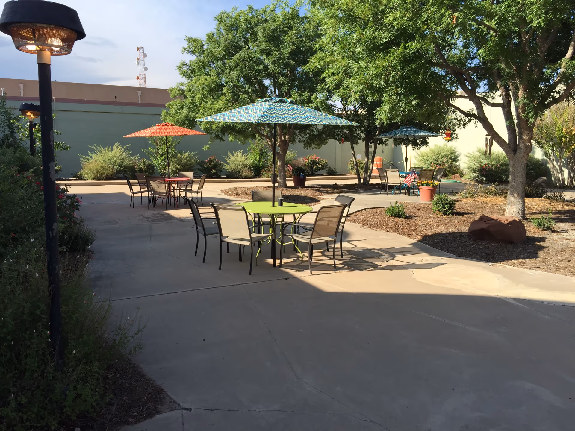 Outdoor patio area with several round tables and chairs under colorful umbrellas. The area is surrounded by trees, plants, and shrubs, with a paved walkway winding through the space. There are potted plants and American flags near some tables, and a tall lamp post is visible on the left side.