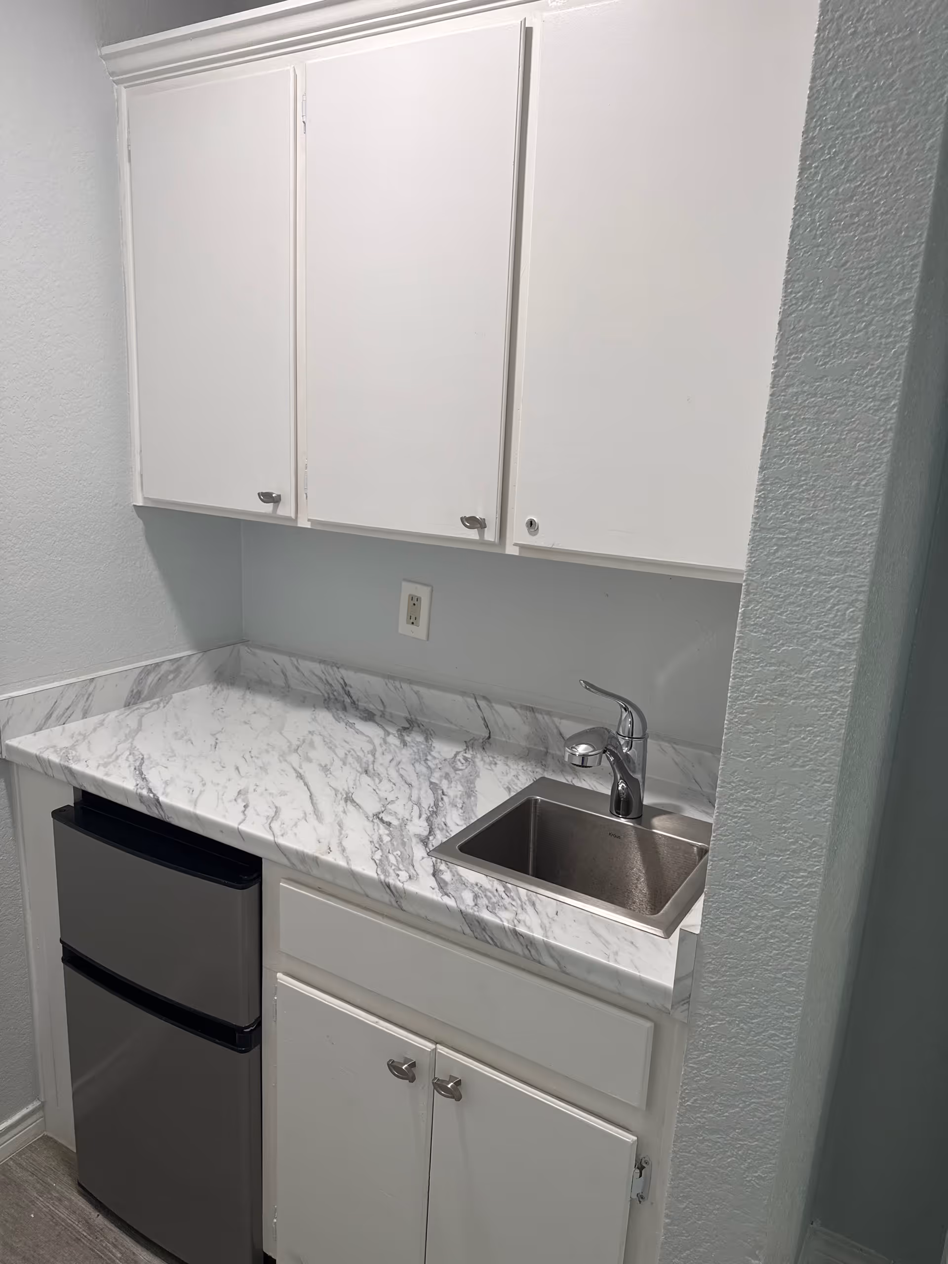 Compact kitchenette with white cabinets, marble-look countertop, and a stainless steel sink.