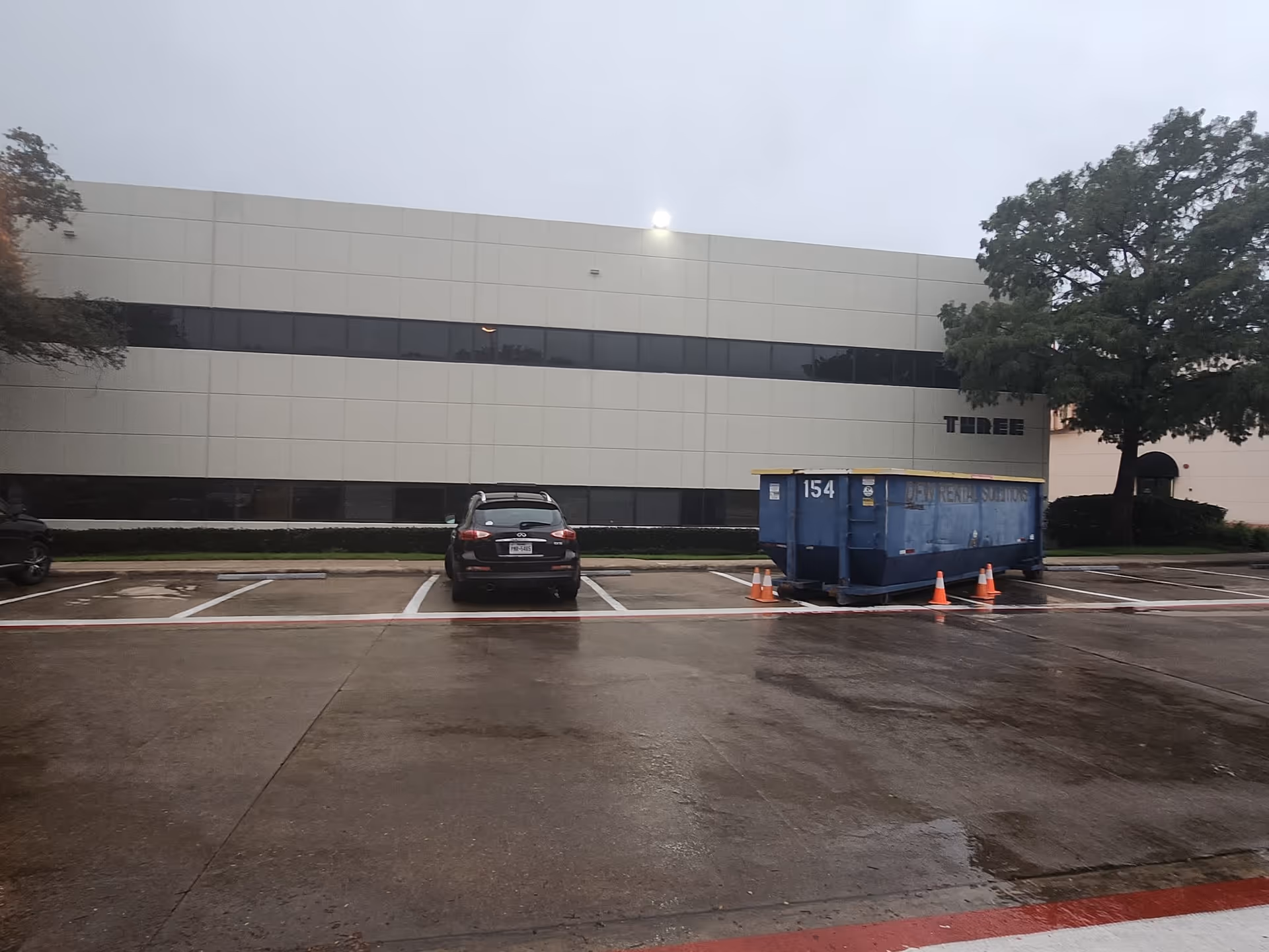 Gray commercial building with a parking lot, black SUV, and blue dumpster.