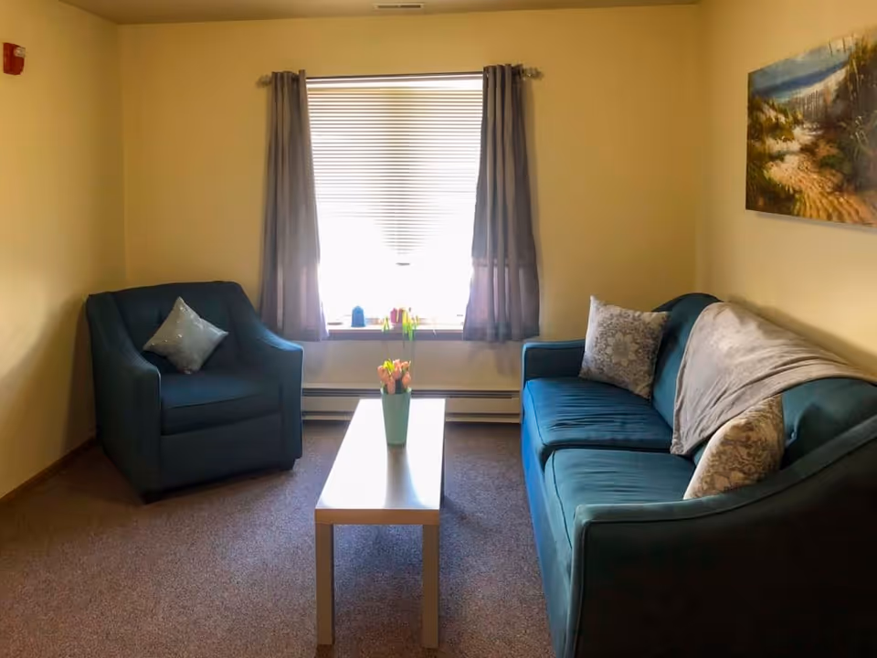 Small living room with a teal armchair and sofa, a coffee table holding a vase of flowers, and a window with curtains.