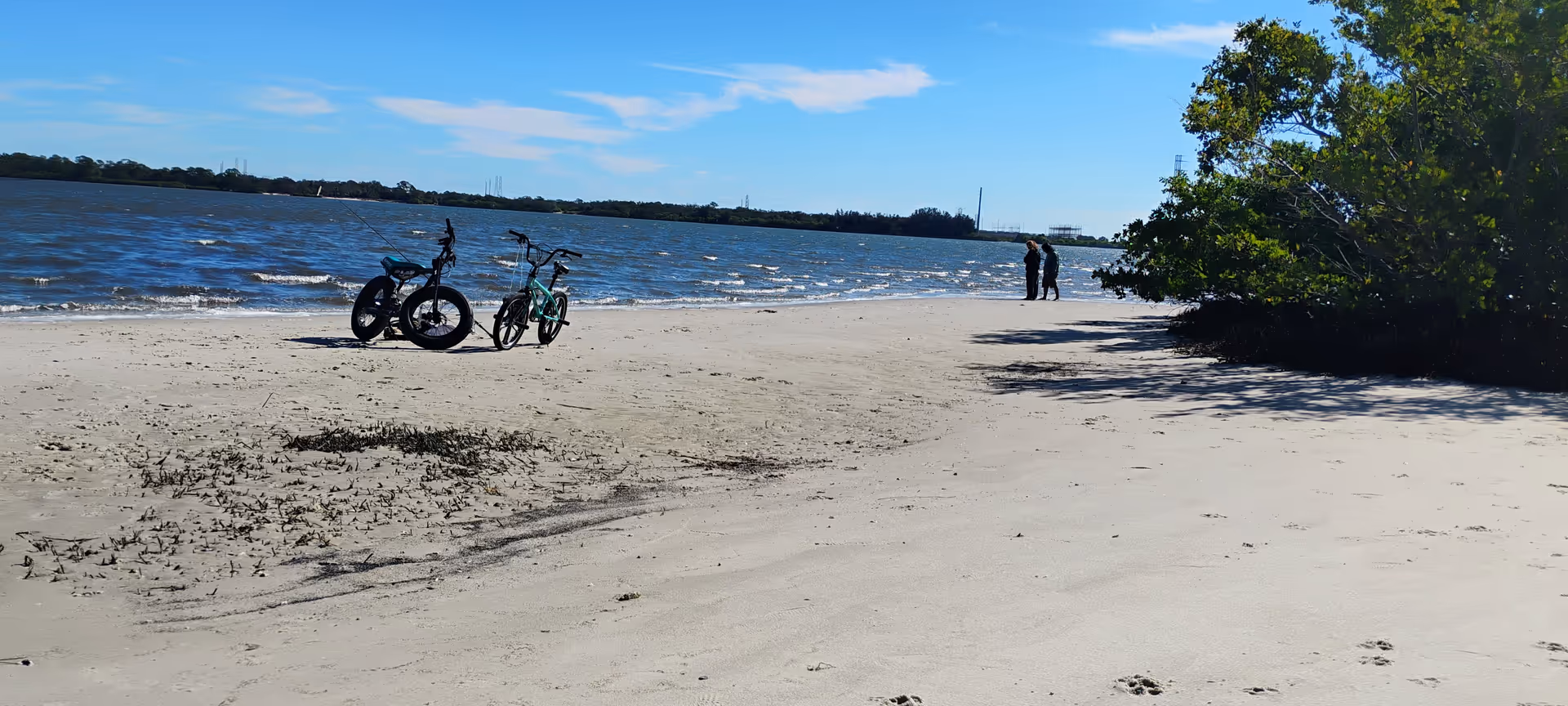 Two bicycles on a sandy beach with calm water and two people walking nearby.