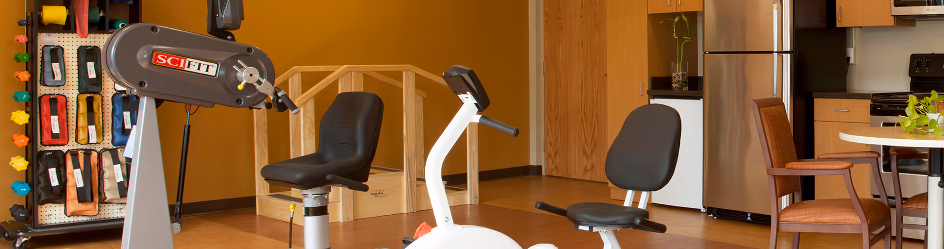 Bright interior rehab area with exercise machines, a wall-mounted resistance bands rack, and a kitchen.
