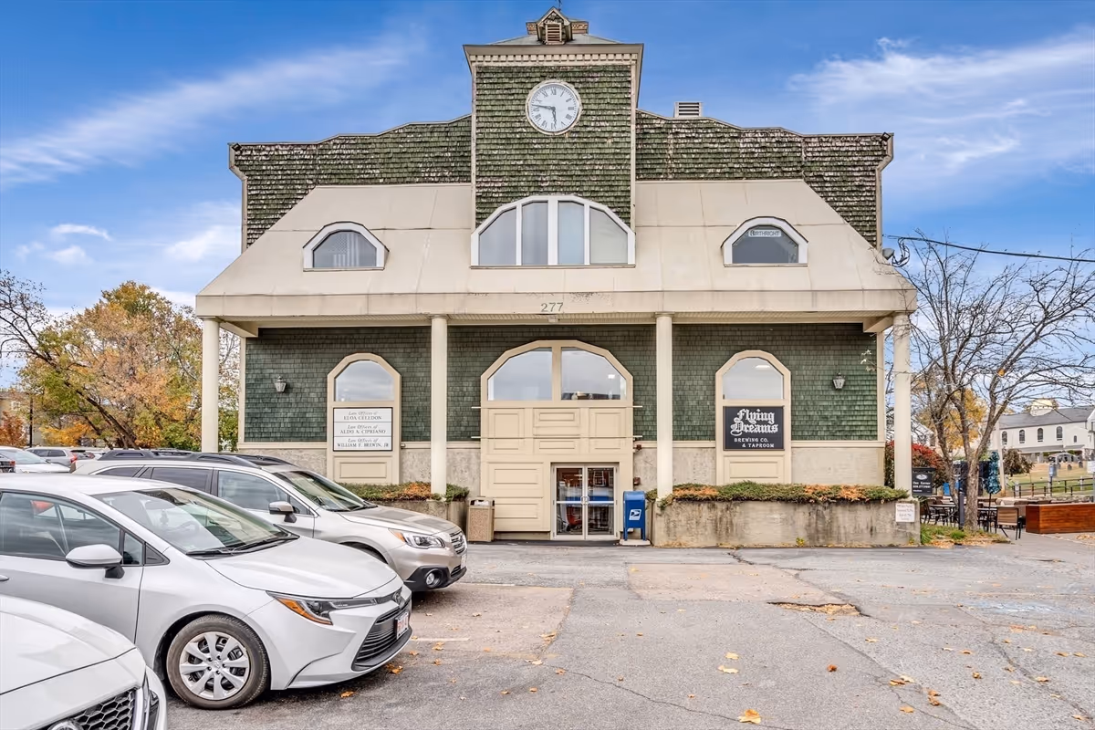 Front view of a green-brick building with clock tower and parked cars.