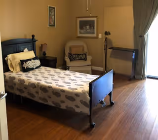 A cozy bedroom in a senior living facility with a bed, chair, and large window.