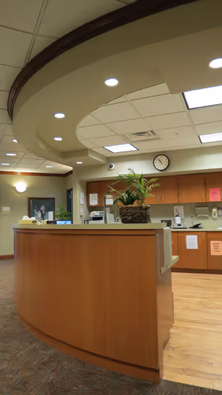 Curved wooden reception desk in a hospital-like lobby with plant and cabinetry.