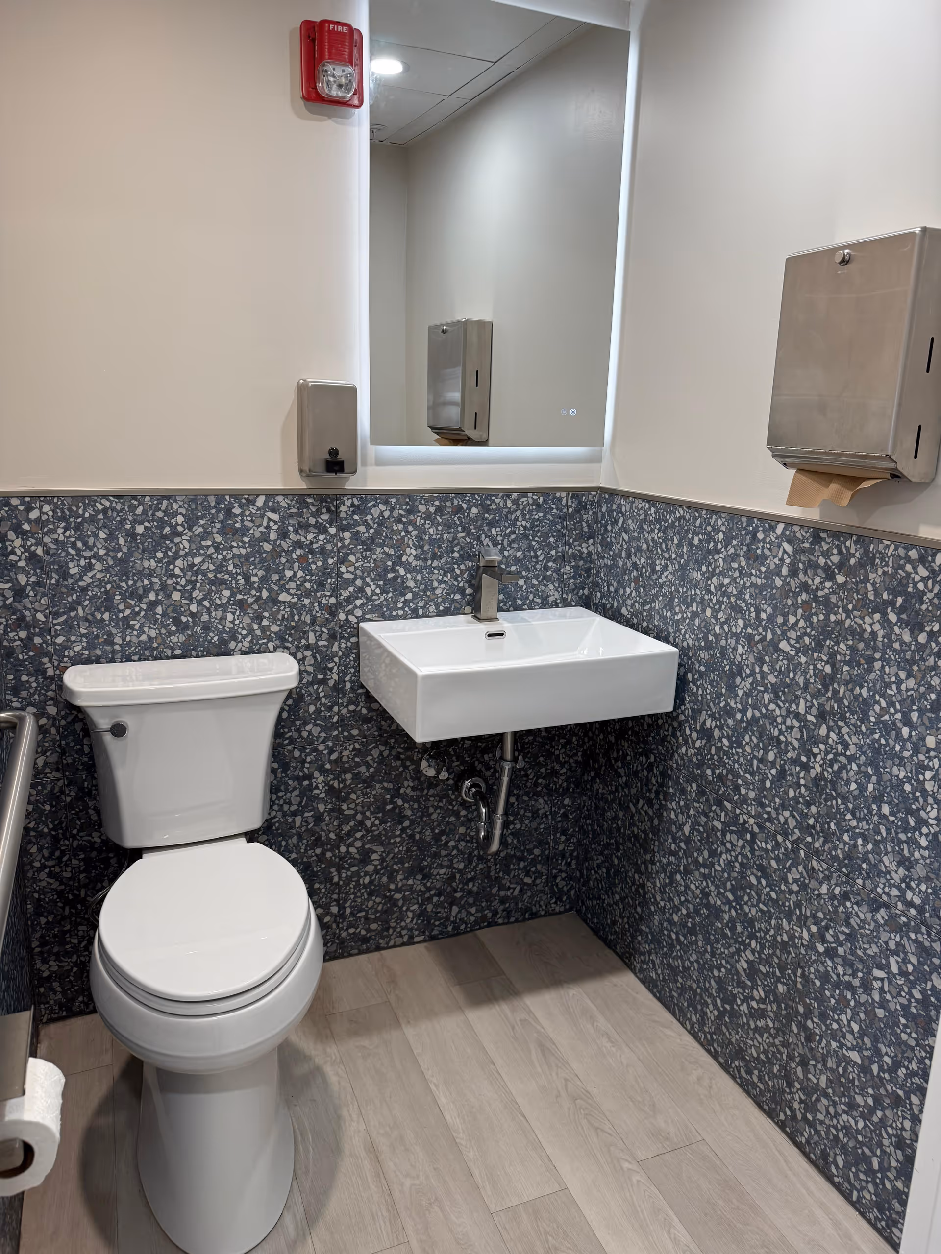 Small bathroom with toilet, wall-mounted sink, mirror, and paper towel dispenser.