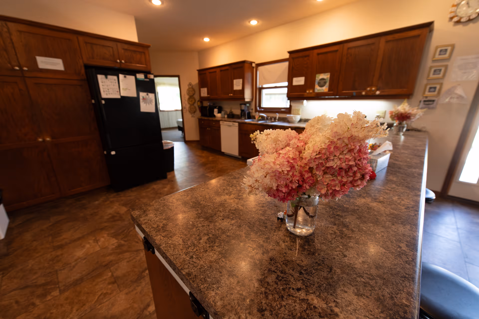 Interior view of a kitchen with brown wooden cabinets, a black refrigerator, a dishwasher, and a countertop with a vase of pink and white flowers. The kitchen has tiled flooring and recessed ceiling lights. There is a window above the sink and a door to the right letting in natural light.