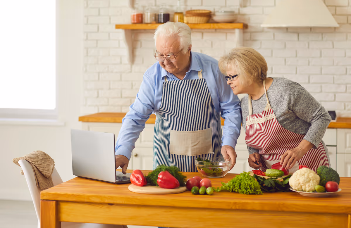 An elderly couple cooks together at a wooden kitchen island with fresh vegetables and a laptop.