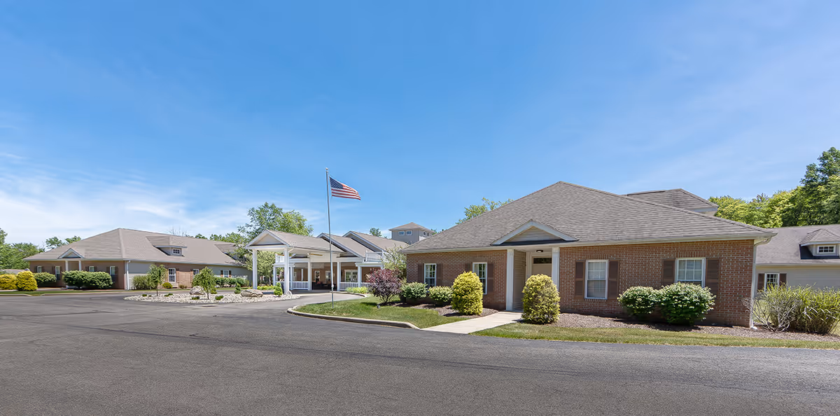 Front exterior of a single-story brick assisted living building with a covered entrance, flagpole, landscaping, and paved driveway.