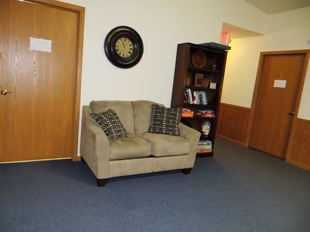A beige loveseat with two pillows sits in a blue-carpeted lounge with a clock and bookshelf.
