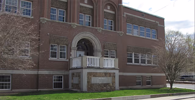 Brick senior living facility with arched entrance and white balcony, grassy lawn.