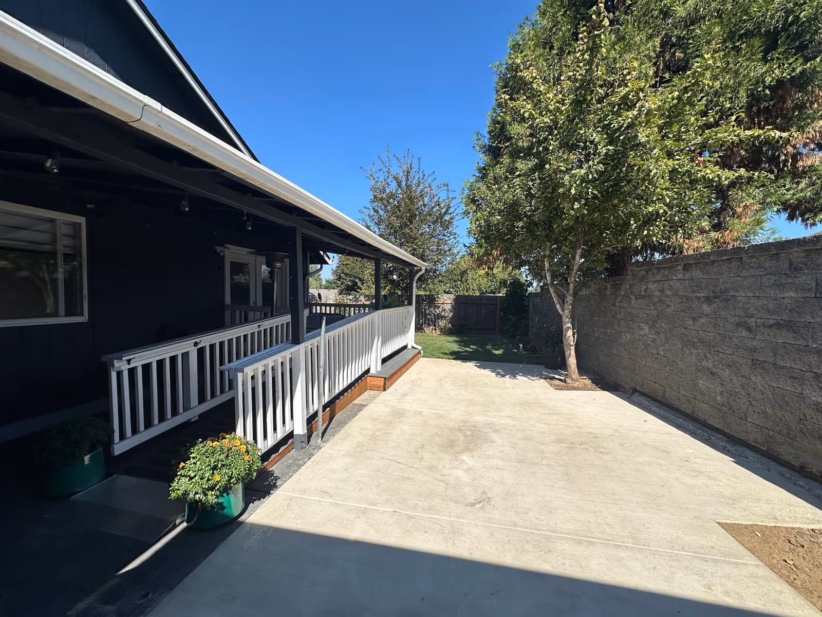 Dark blue single-story building with white railing porch, concrete yard, and trees by a stone wall.