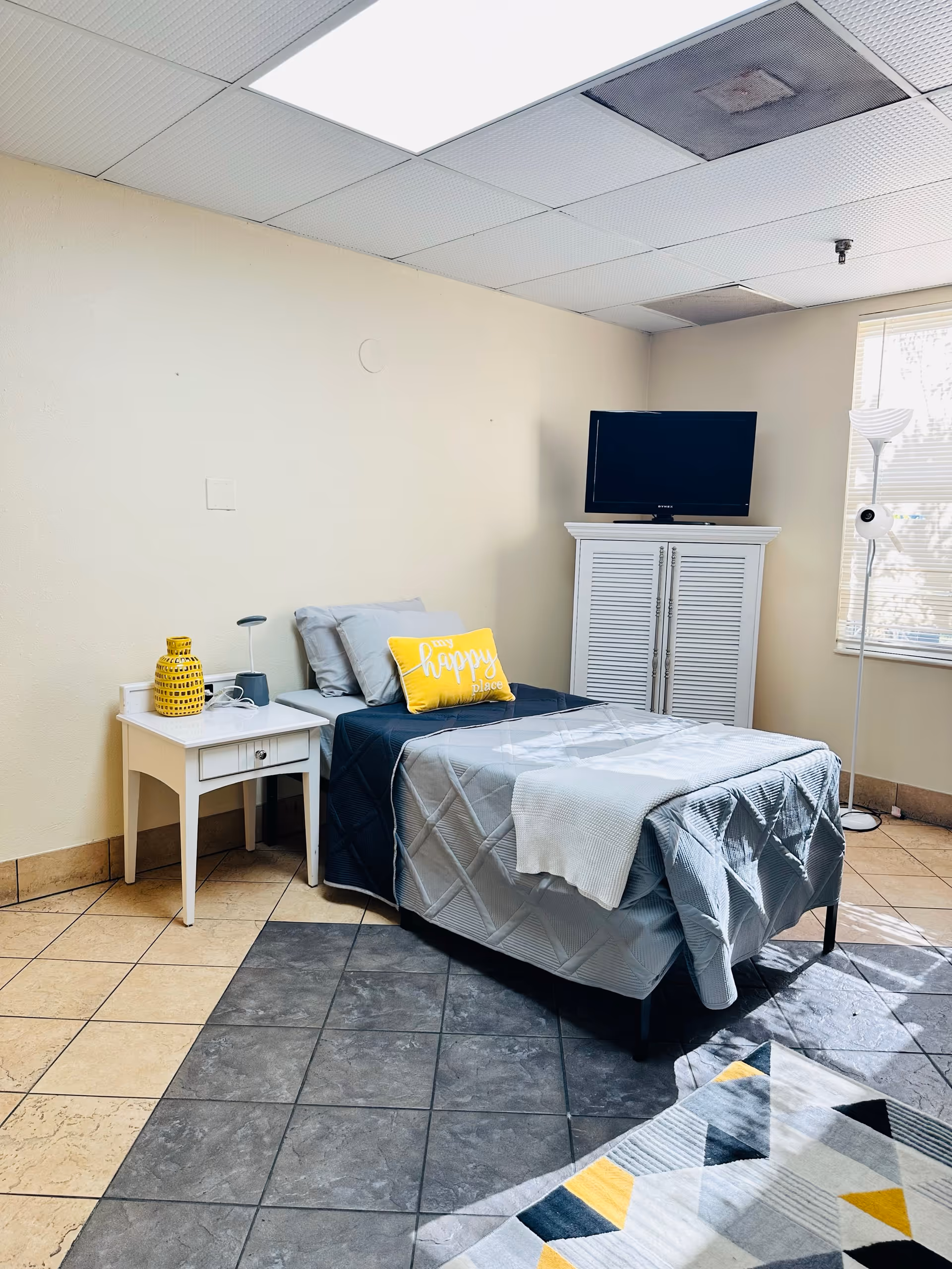 A small, neatly arranged bedroom with a single bed covered in gray and navy blue bedding, a yellow pillow with the words 'happy place', a white nightstand with a yellow vase and a small lamp, a white cabinet with a flat-screen TV on top, a standing lamp near a window with blinds, and a patterned rug on the tiled floor.