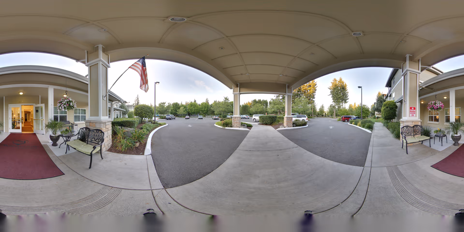 Covered entrance area of Woodland by Bonaventure senior living facility with benches, potted plants, hanging flower baskets, an American flag, and a view of the parking lot and surrounding greenery.