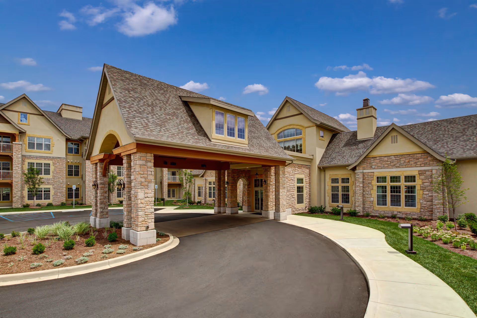 Exterior view of The Plaza at The Barrington of Carmel senior living facility showing a covered entrance with stone pillars, a curved driveway, landscaped greenery, and a multi-story building under a blue sky with scattered clouds.