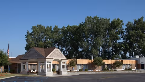 Exterior view of Medilodge of Lansing facility showing a single-story building with a covered entrance, surrounded by trees and a clear blue sky.
