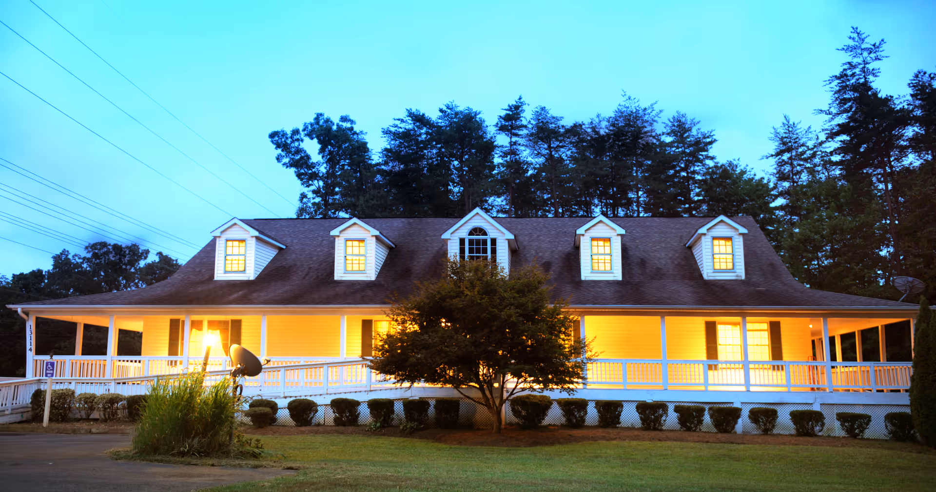 Exterior view of a large, two-story house with a wrap-around porch illuminated by warm lights at dusk. The house has multiple dormer windows on the roof and is surrounded by trees and neatly trimmed bushes. A tree stands in front of the porch, and there is a satellite dish mounted near the porch.