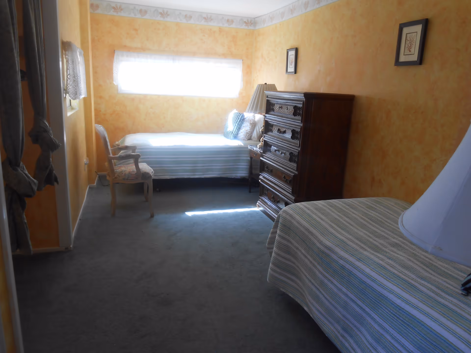 A small bedroom with two single beds covered in striped bedspreads, a wooden chest of drawers, a wooden chair with floral upholstery, and a window with sheer white curtains letting in bright light. The walls are painted in a warm yellow-orange tone with a floral border near the ceiling.
