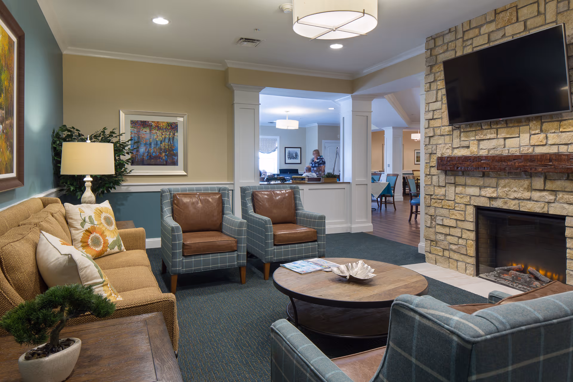 A cozy senior living facility common area with a stone fireplace and a mounted flat-screen TV above it. The room features a beige sofa with floral pillows, two plaid armchairs with brown leather cushions, a round wooden coffee table with a decorative bowl and magazines, a table lamp, framed artwork on the walls, and a potted plant. In the background, there is a dining area with tables and chairs, and a person standing near the far wall.