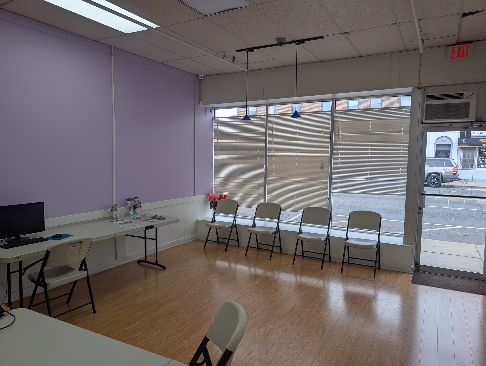 Lavender-walled interior room with a long desk, computer, and folding chairs by windows.