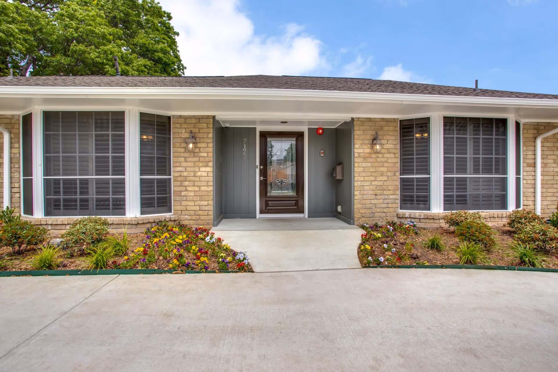Front entrance of a single-story brick building with a glass door and two large bay windows on either side. There are flower beds with colorful flowers and shrubs along the walkway leading to the door. The sky is partly cloudy with some blue visible.