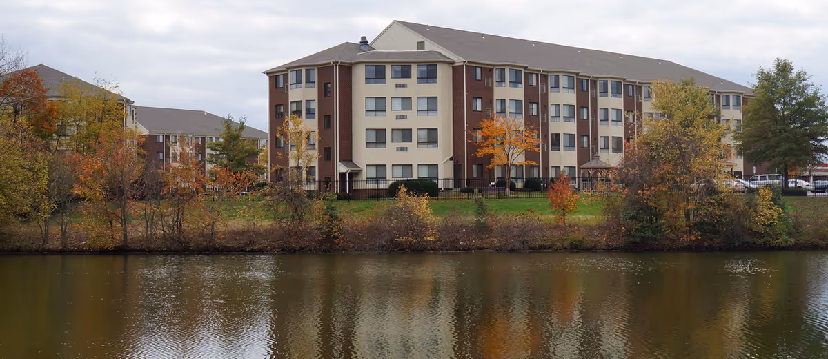 A multi-story senior living building sits along a river with autumn-colored trees.