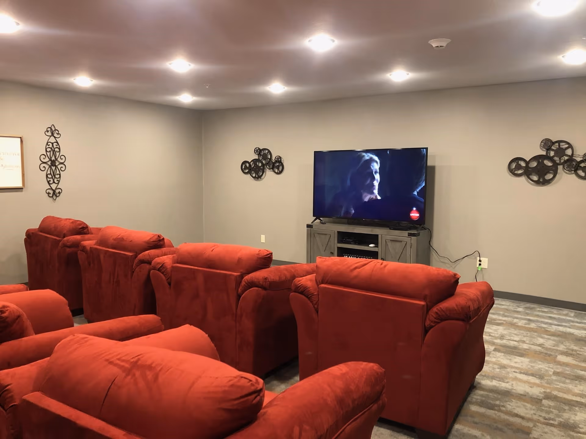 A cozy living room with multiple red upholstered armchairs arranged in rows facing a flat-screen TV mounted on a gray wooden stand. The walls are painted light gray and decorated with metal wall art. The floor has a wood-like pattern, and the ceiling has recessed lighting.