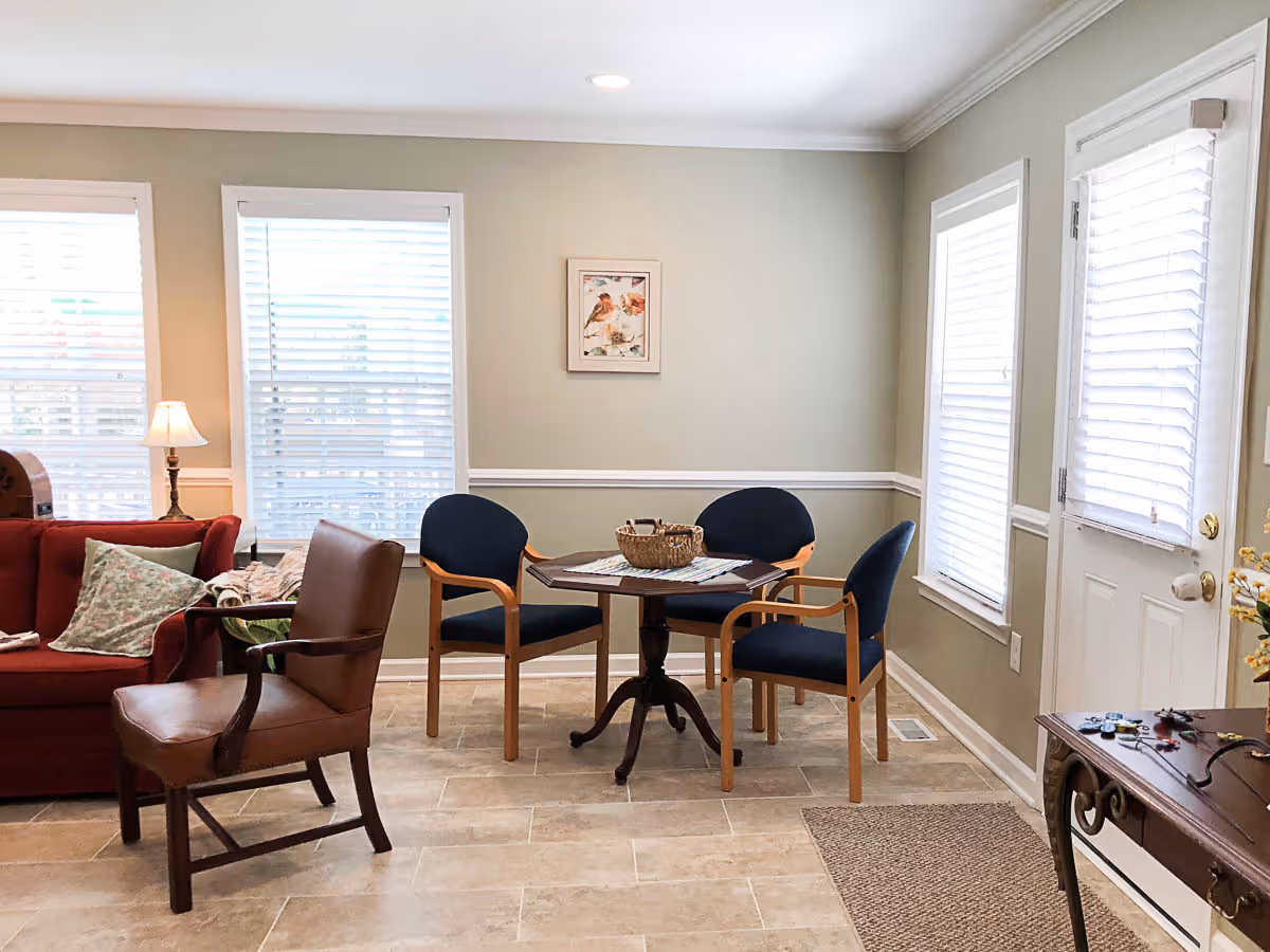 A cozy living room area with a red sofa, a brown leather armchair, and a small round table surrounded by three blue cushioned chairs. The room has beige tiled flooring, light gray walls with white trim, three windows with white blinds, a white door with a window, and a small table with decorative items near the door.