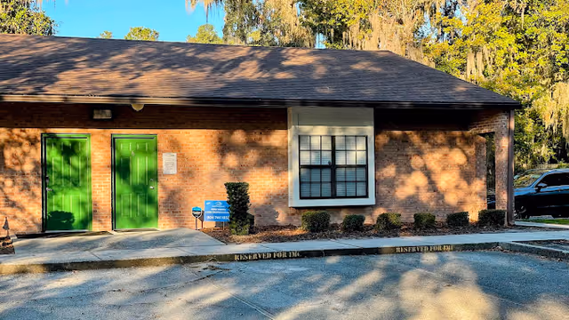 Brick facility front with green doors, window, shrubs, and a parking lot.