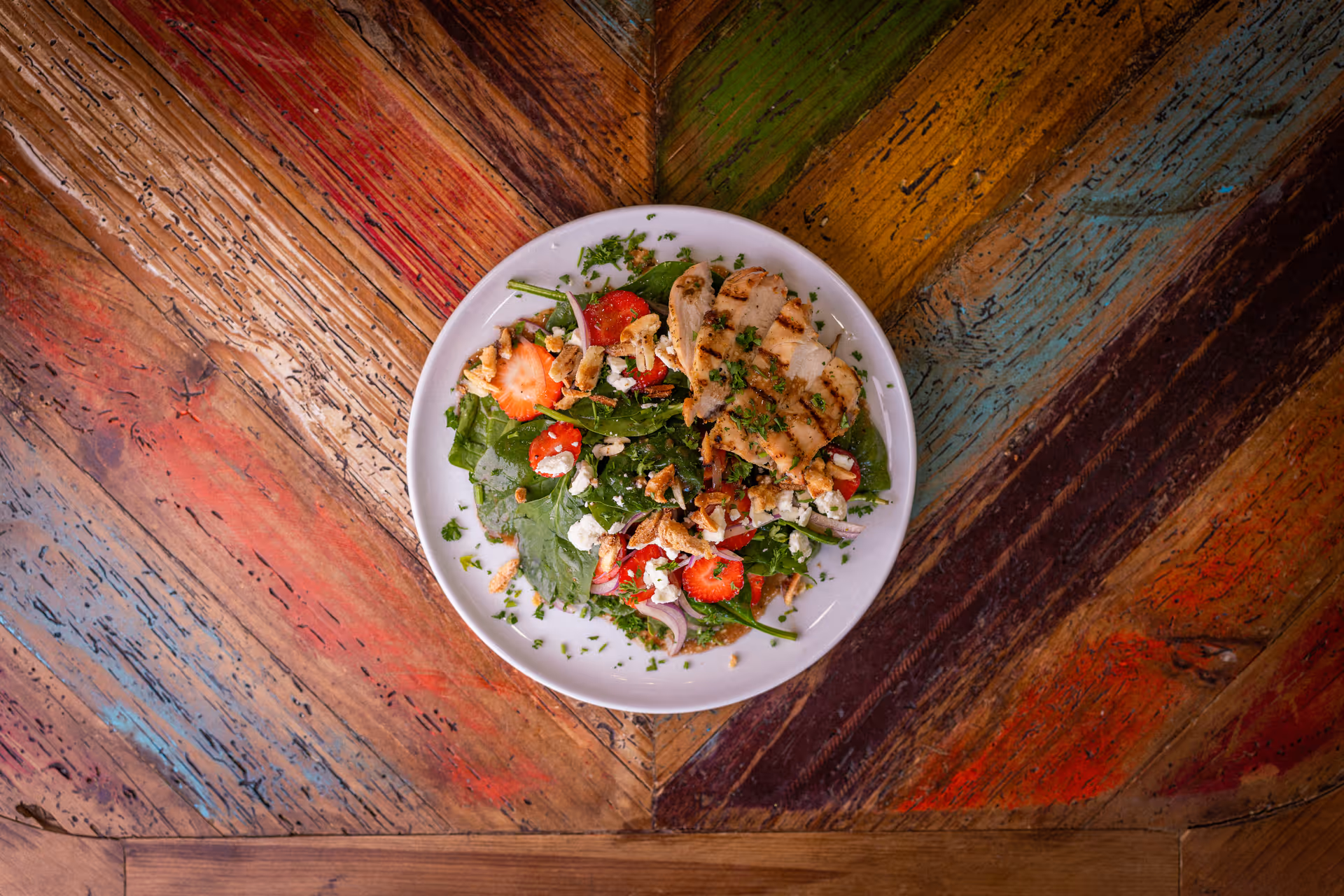 A white plate with a fresh salad consisting of grilled chicken, spinach leaves, sliced strawberries, walnuts, red onions, and crumbled cheese, placed on a colorful rustic wooden table.
