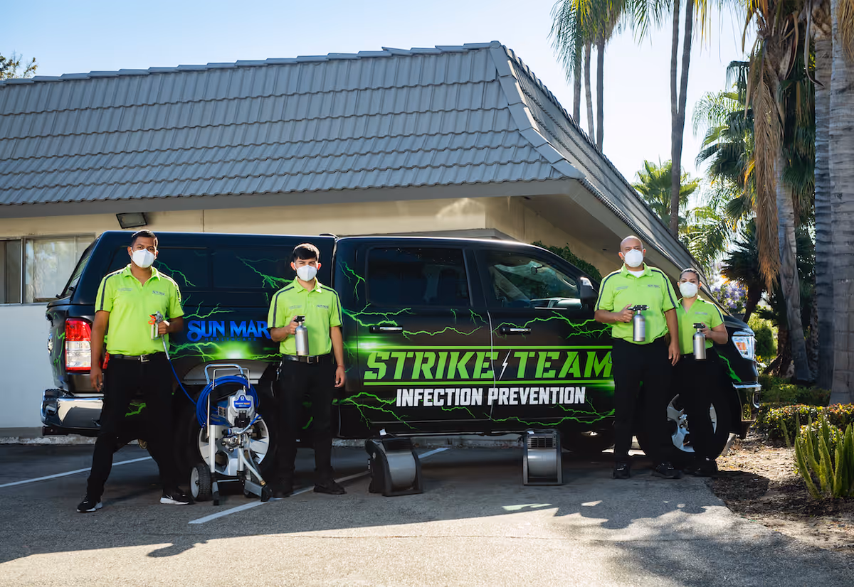 Four people wearing bright green shirts and face masks stand in front of a black vehicle with green lightning graphics and the text 'STRIKE TEAM INFECTION PREVENTION'. They are holding spray bottles and standing next to cleaning equipment in a parking lot outside a building with palm trees in the background.
