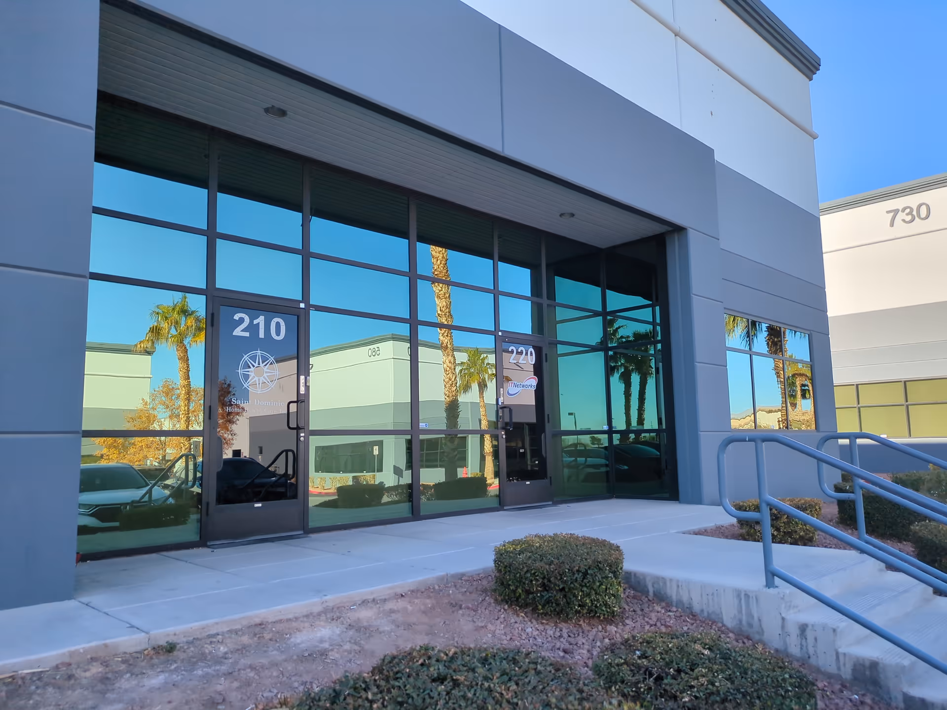 Modern glass-front entrance with doors labeled 210 and 220, ramp, desert landscaping.
