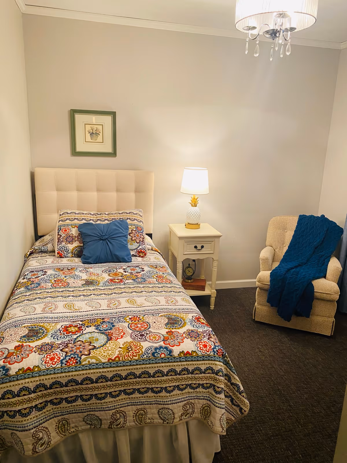 A cozy bedroom with a single bed covered in a colorful floral and paisley patterned bedspread. There is a blue decorative pillow on the bed. Next to the bed is a white nightstand with a pineapple-shaped lamp and a small clock on the lower shelf. A beige armchair with a blue knitted throw blanket draped over it is positioned in the corner. A framed picture hangs on the wall above the bed, and a chandelier-style light fixture is visible on the ceiling.