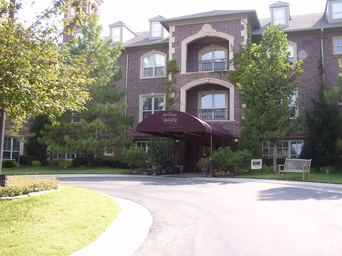 Front exterior view of a three-story brick building with a maroon canopy entrance labeled 'The Mansion at Waterford'. The building is surrounded by green trees and shrubs, with a curved driveway leading to the entrance. There is a wooden bench and a small sign on the lawn near the entrance.