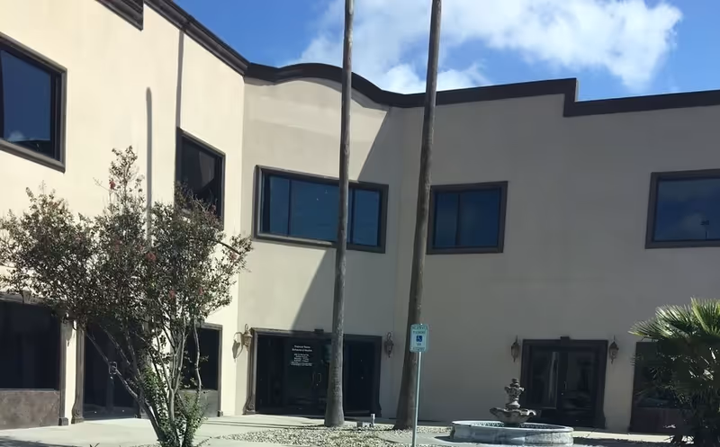 Beige facility exterior with large windows, palm trees, and a fountain in the courtyard.