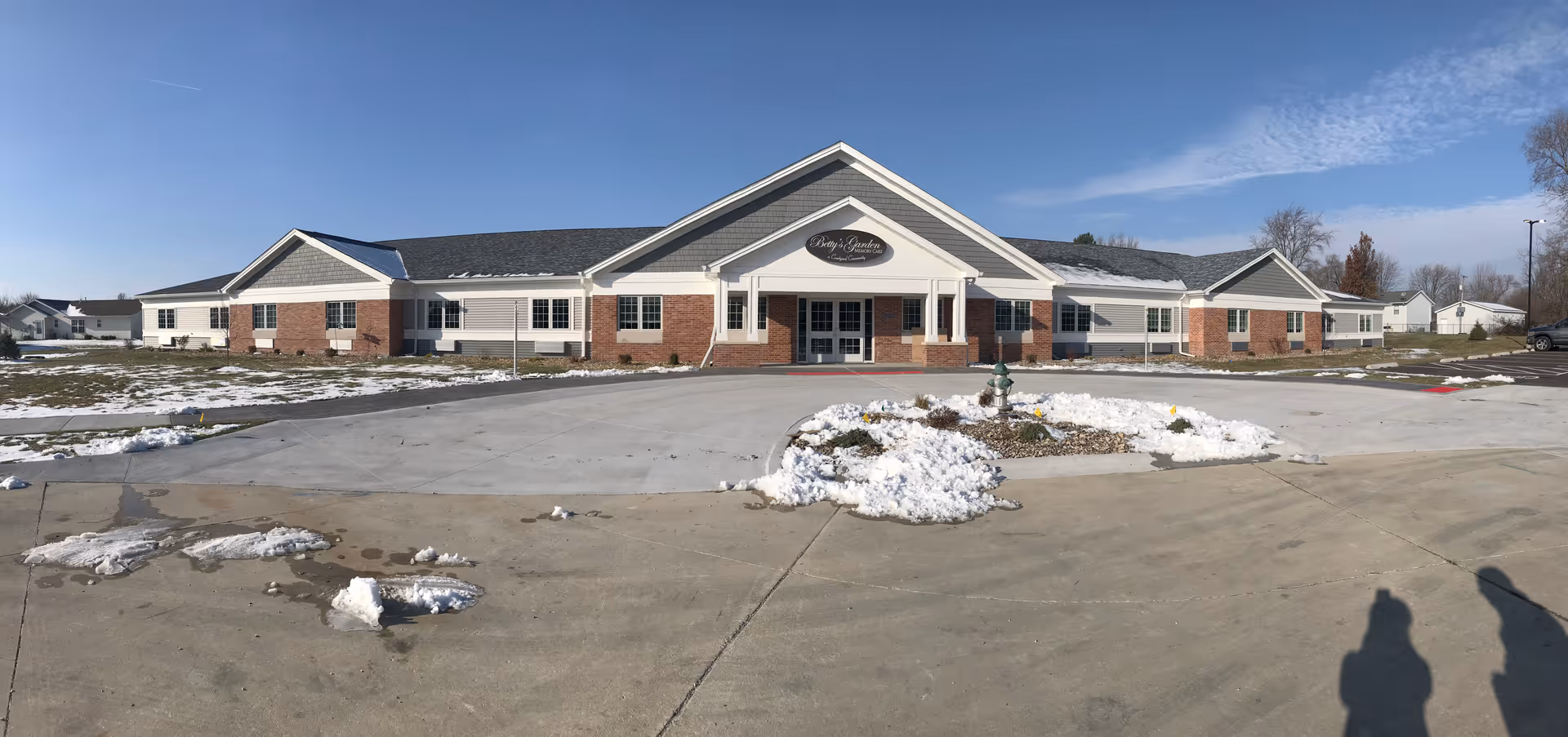 Front exterior view of a single-story senior living facility building with a combination of brick and siding, a peaked roof, and a covered entrance. There is some snow on the ground and a clear blue sky above.