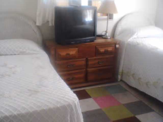Two twin beds with white bedspreads flank a wooden dresser topped by a TV and lamp in a small bedroom.