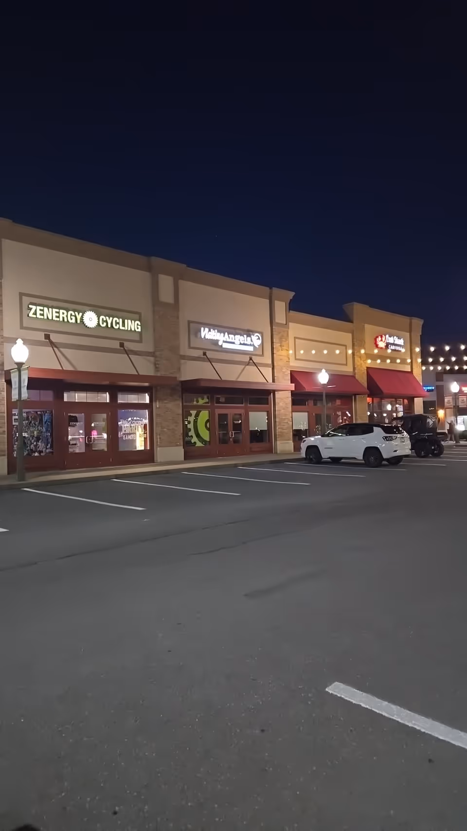 Night exterior of a shopping plaza with a Visiting Angels storefront sign.