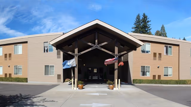 Exterior view of a senior living facility building with a covered entrance. The building is two stories tall with beige siding and multiple windows. Two flags, one American and one light blue, hang from the covered entrance. There are potted plants near the entrance and a clear blue sky above.