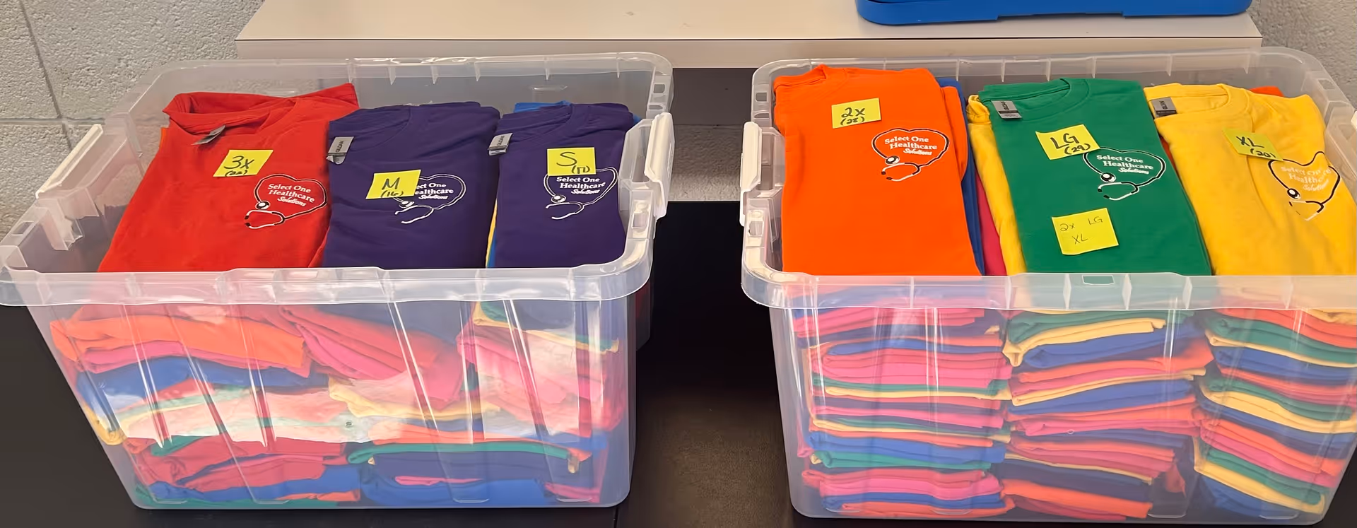 Clear plastic bins hold folded, colorful shirts labeled by size on a table.